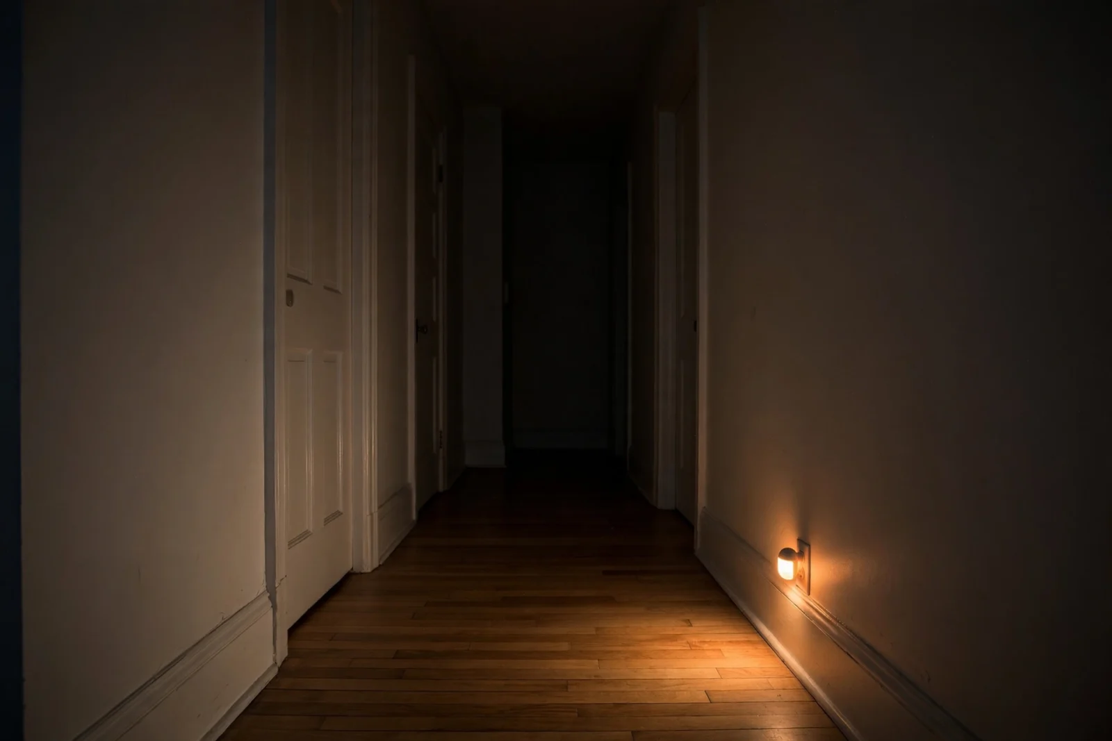 A dim hallway at night lit by the warm glow of a plug-in motion-activated night light near the baseboard.