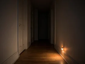 A dim hallway at night lit by the warm glow of a plug-in motion-activated night light near the baseboard.